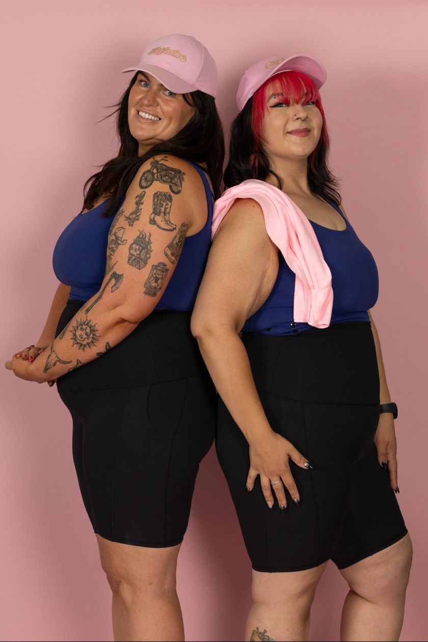 Two women wearing matching blue tops and black shorts with pink visors against a pink background.